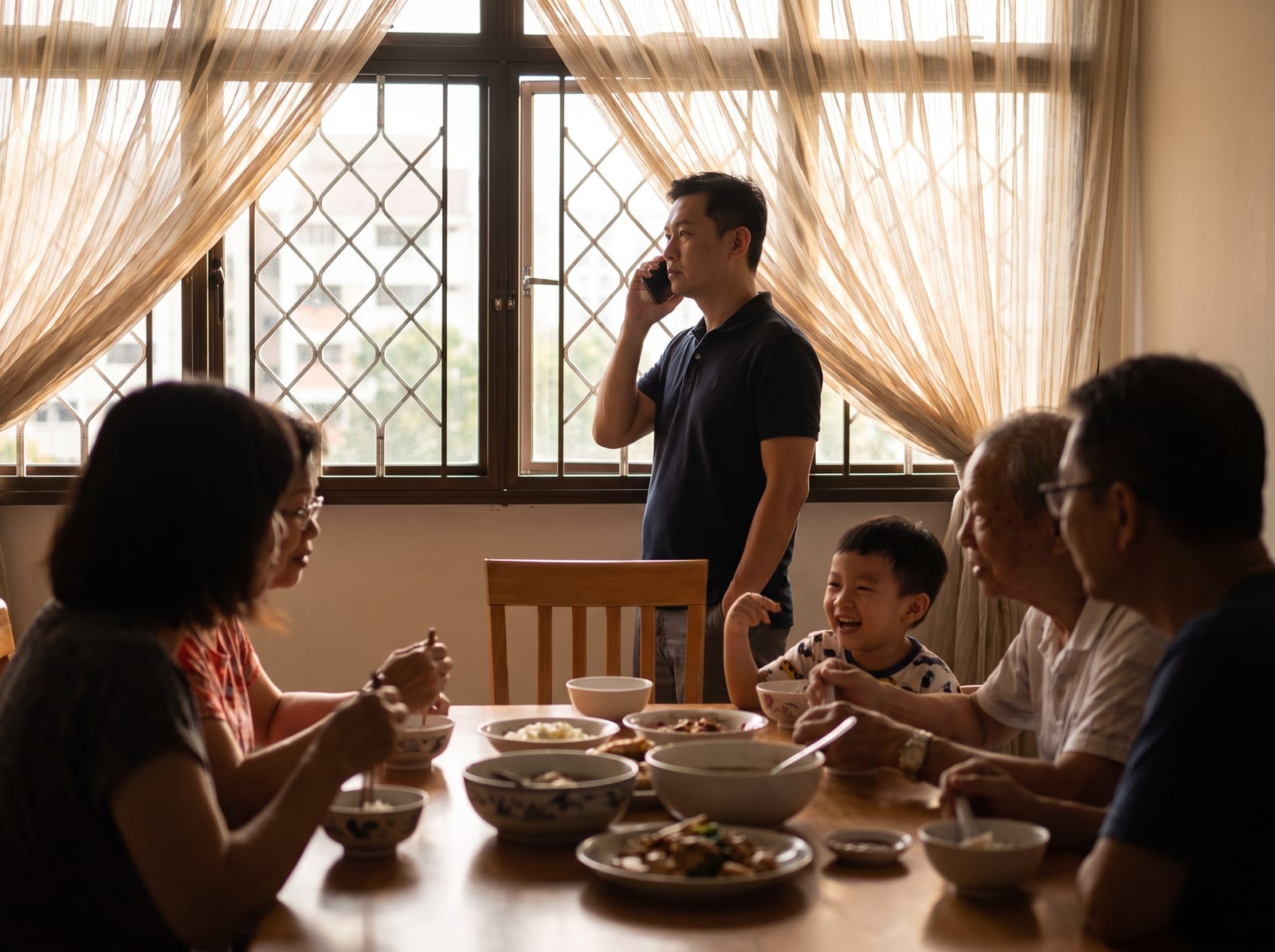A Sunday family dinner in a Singapore flat — the father stands apart at the window, on a work call.